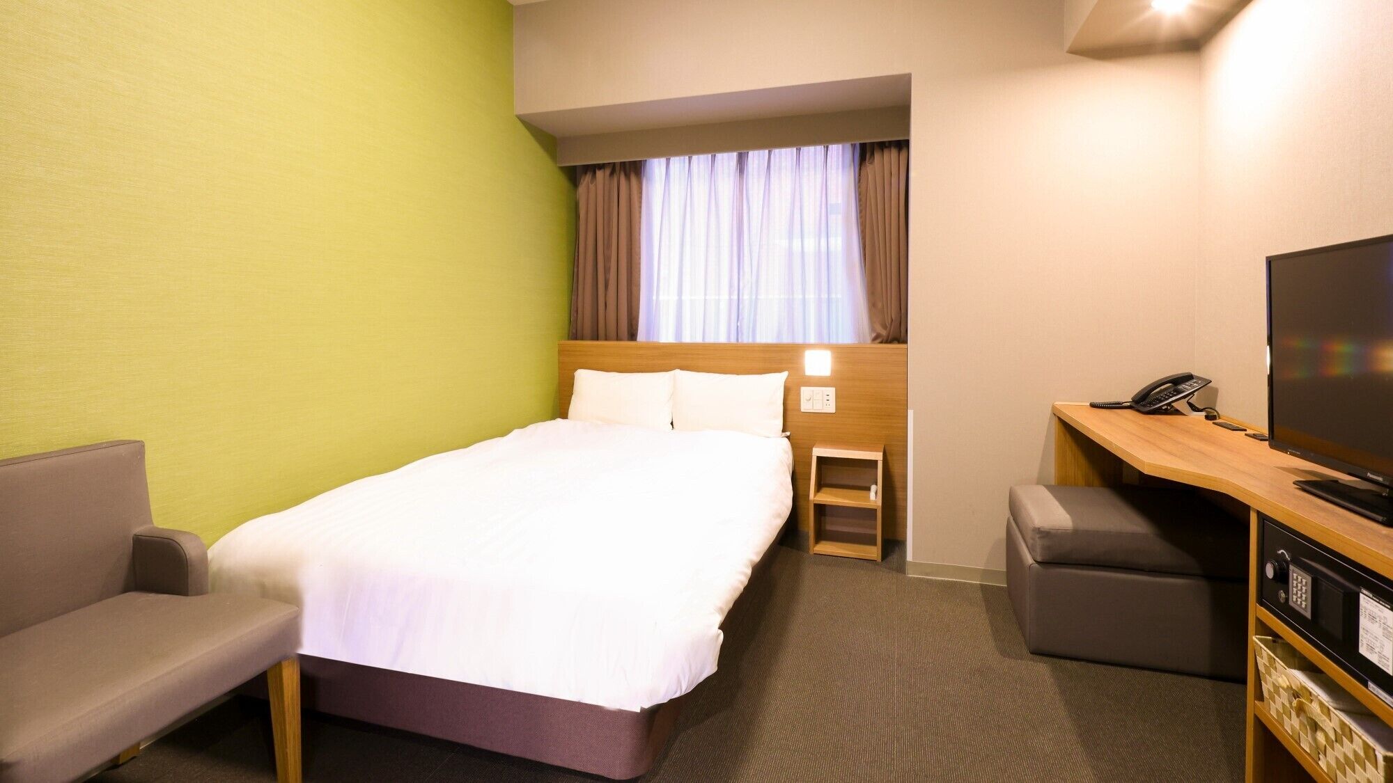 A hotel room with a bed, a desk with a TV, a chair, and a small ottoman.