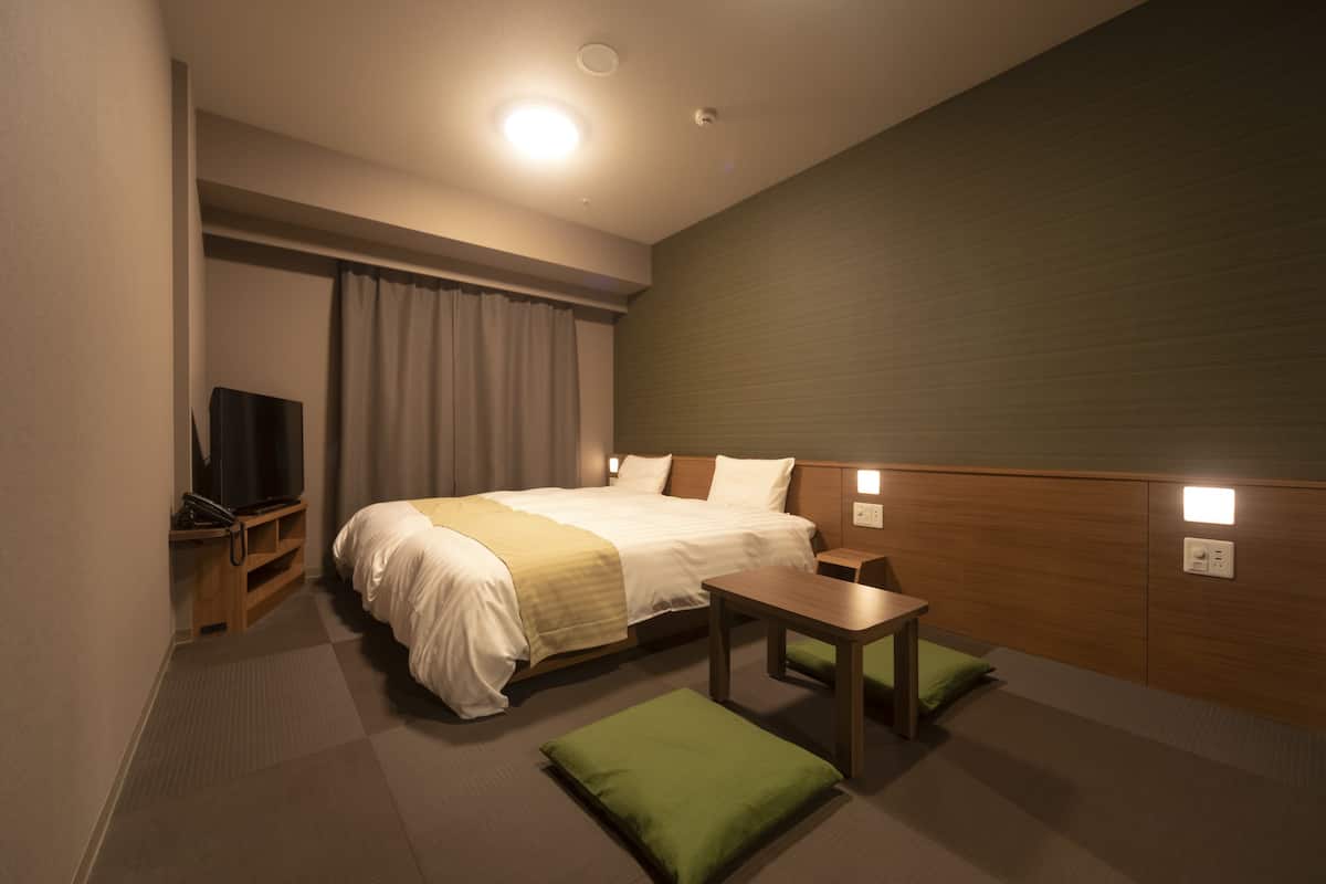 Ein Hotelzimmer mit Bett, Nachttisch, grünen Kissen, Fernseher und Wandleuchte.