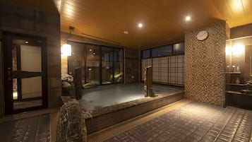 Perinteinen japanilainen onsen-kylpylä, jossa on kivinen kylpyallas, puinen katto ja laatoitetut seinät.