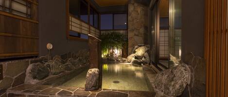 A stone hot spring bath area with a wooden ceiling and large windows.