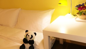 Una cama con ropa de cama blanca, un peluche de panda pequeño, una mesita de noche blanca, una lámpara de escritorio amarilla y una cesta de flores.