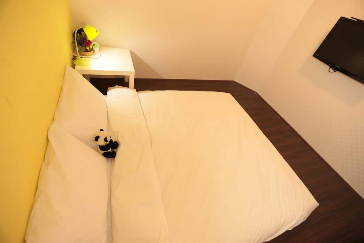 A neatly made bed with white bedding, a small bedside table with a lamp, and a wall-mounted TV.