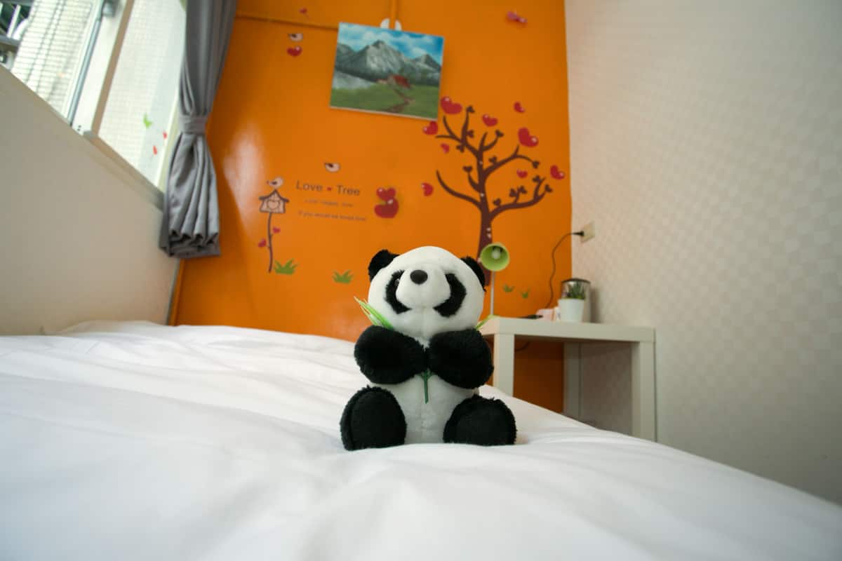 A bed with a stuffed panda toy, a wall with a tree decal, and a small white nightstand with a plant.