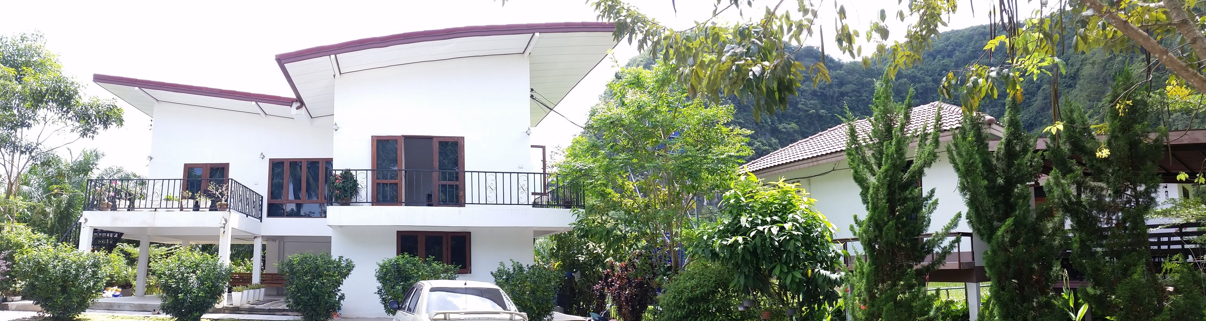 A two-story house with a balcony, surrounded by lush greenery and trees.