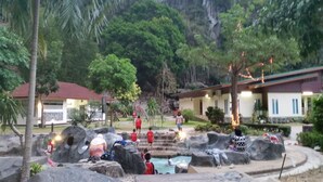 Un área de piscina con rocas y una fuente de agua, rodeada de árboles y un edificio con techo rojo.