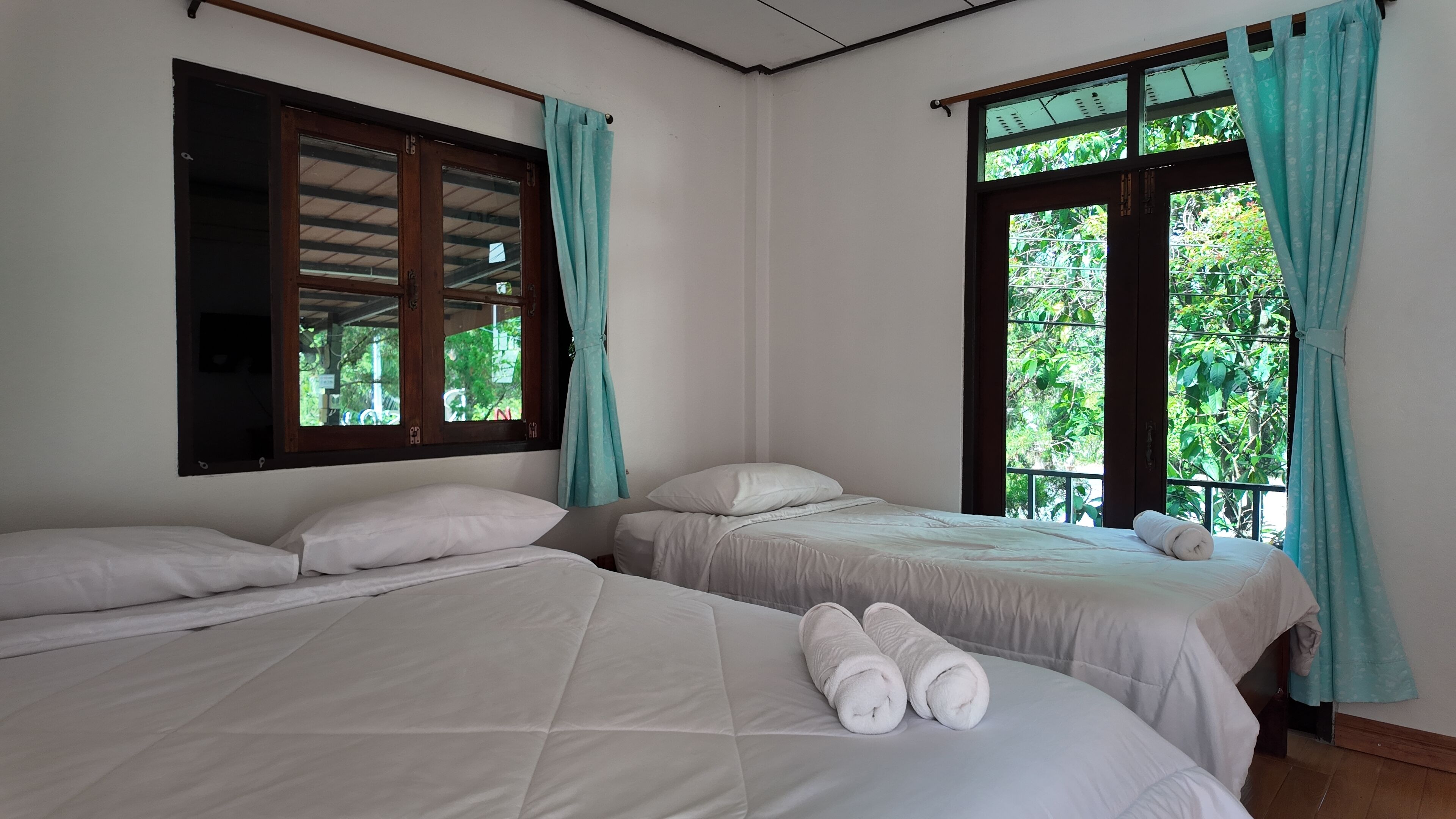 A room with two beds, a window with curtains, and a view of greenery outside.