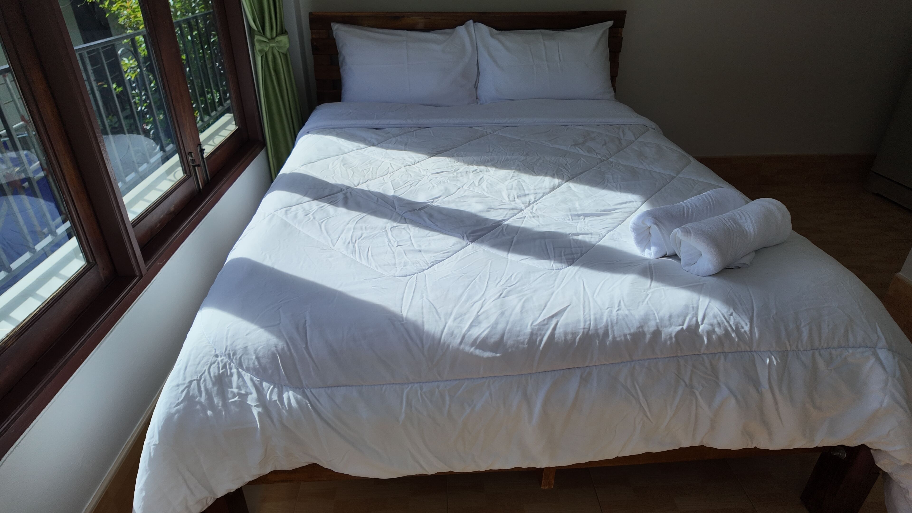 A neatly made bed with white bedding and a rolled towel, positioned next to a window with a view of greenery outside.