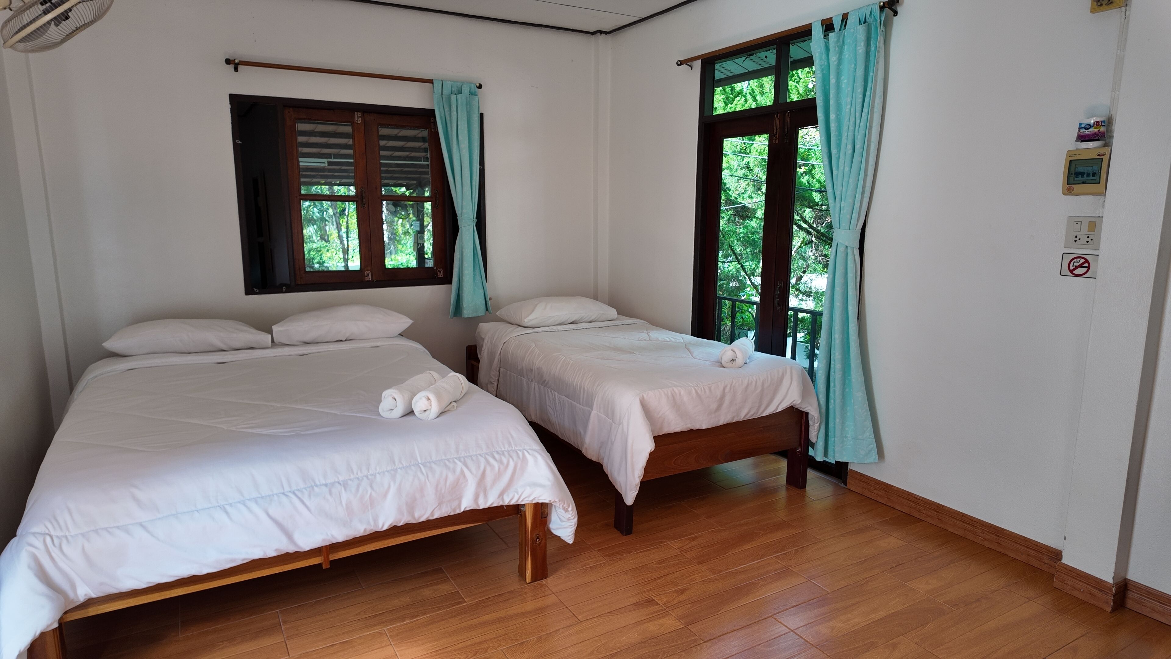 A room with two wooden beds, a window with curtains, a ceiling fan, and a thermostat on the wall.