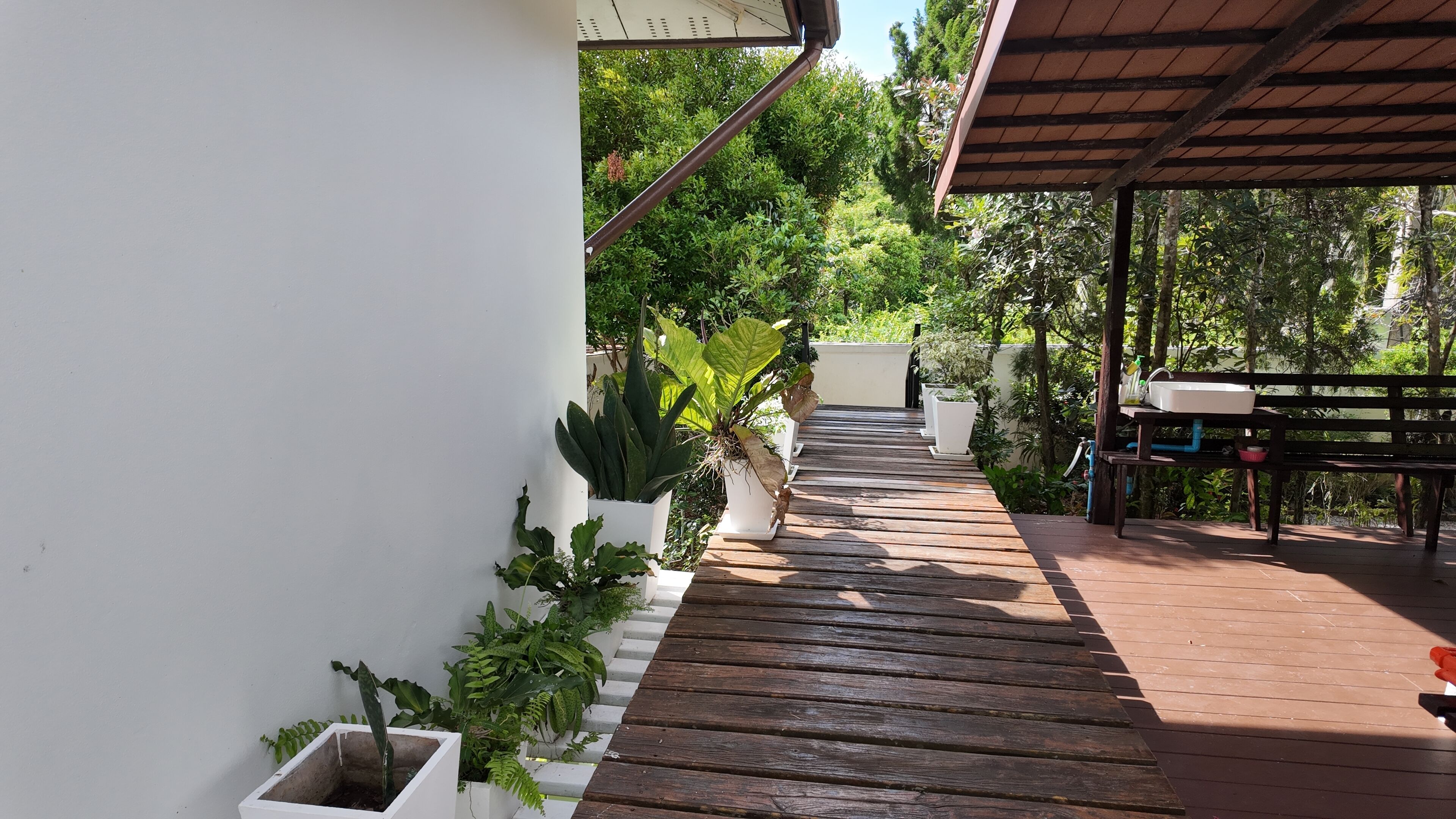 Una terraza de madera con plantas en macetas, una pared blanca y un espacio cubierto con un banco y un lavabo.