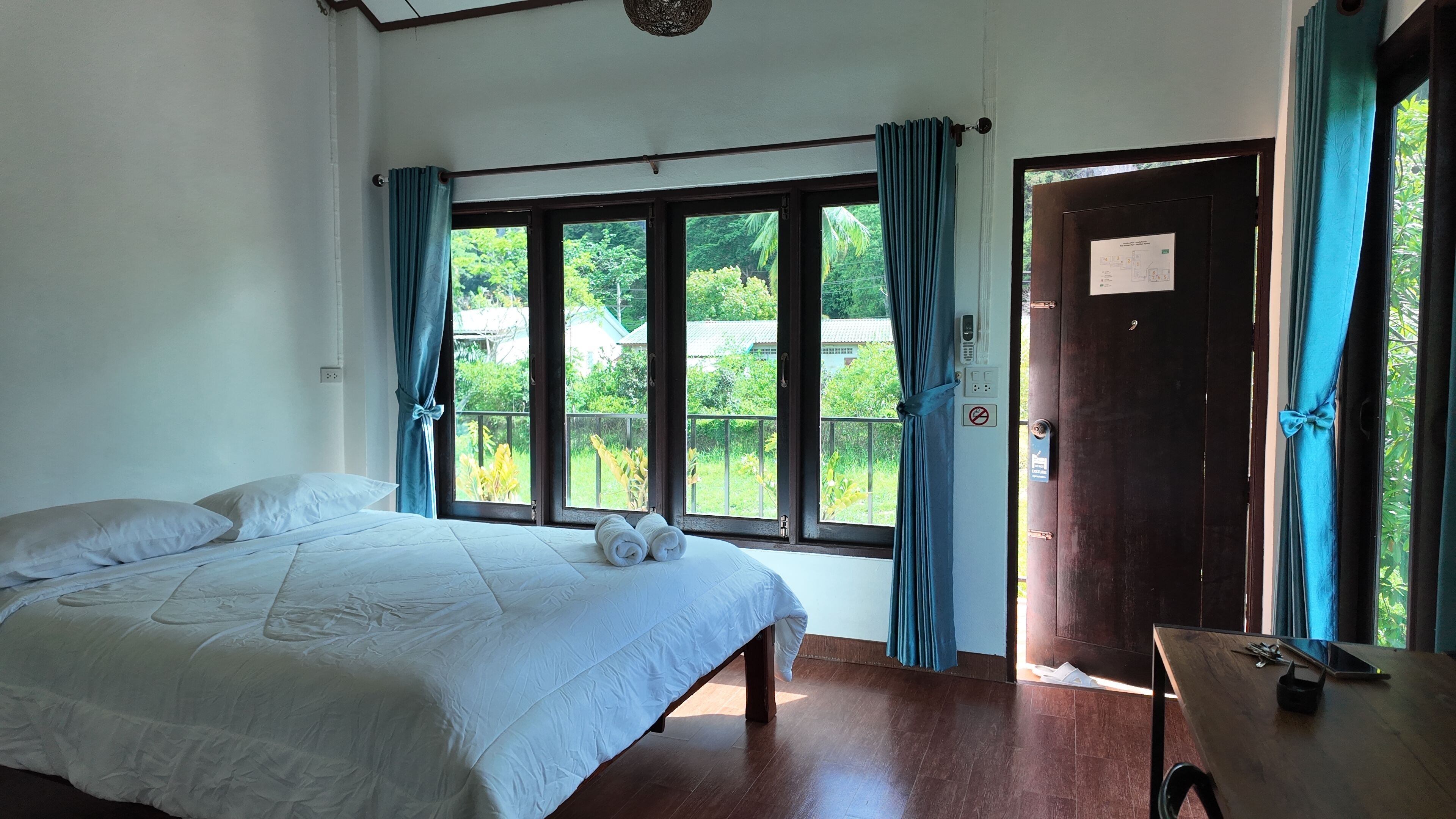 A bedroom with a bed, a wooden table, and a view of greenery through large windows.