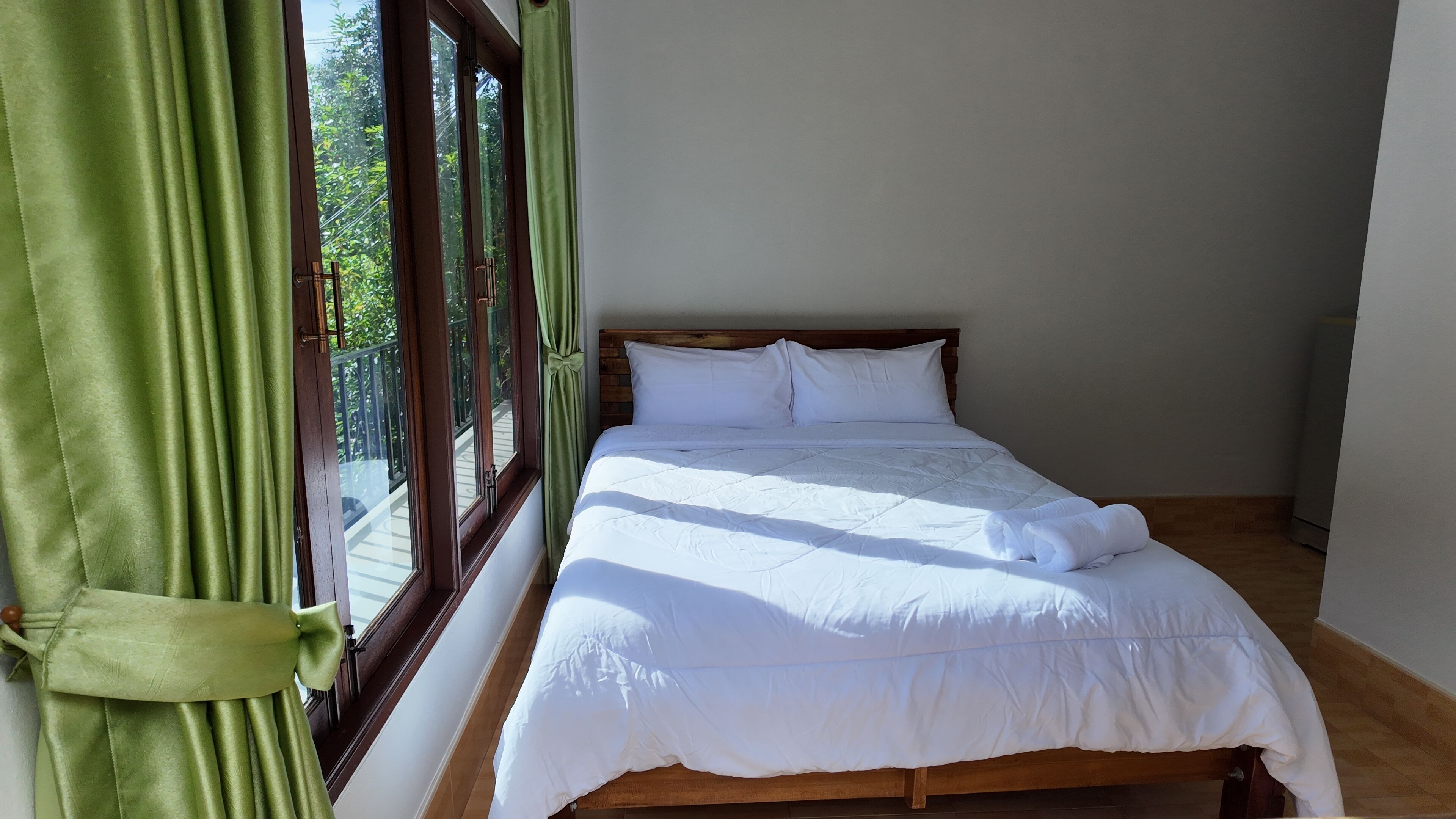 Un lit bien fait, avec du linge de lit blanc, dans une chambre dotée d’une tête de lit en bois et offrant une vue par une fenêtre ornée de rideaux verts.