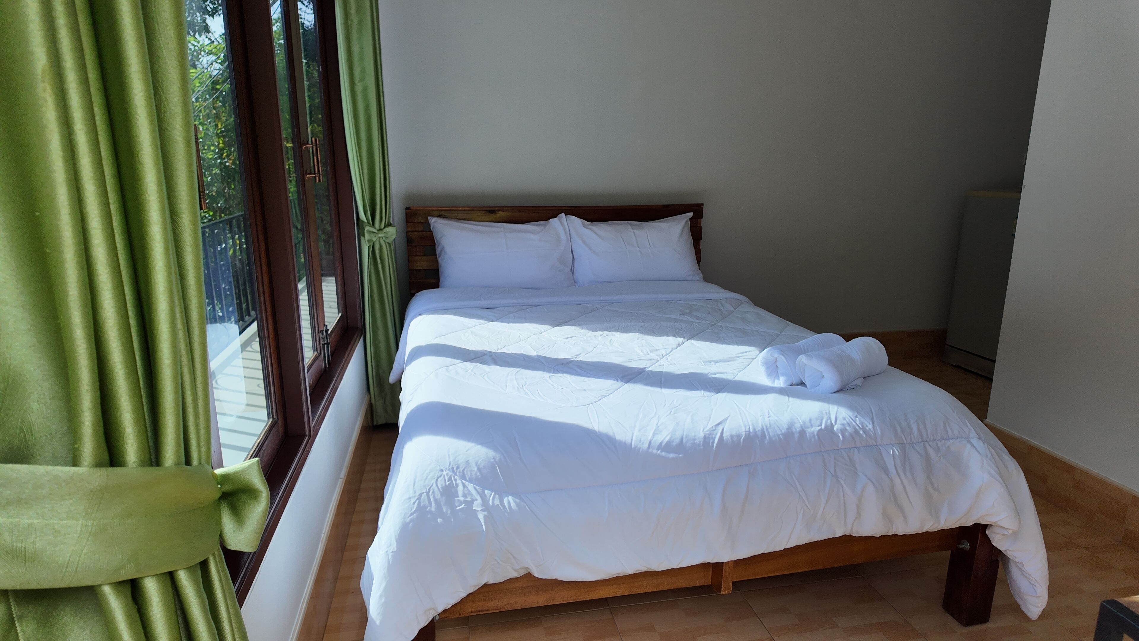 A single bed with a wooden frame and white bedding, a window with green curtains, and a white wall.