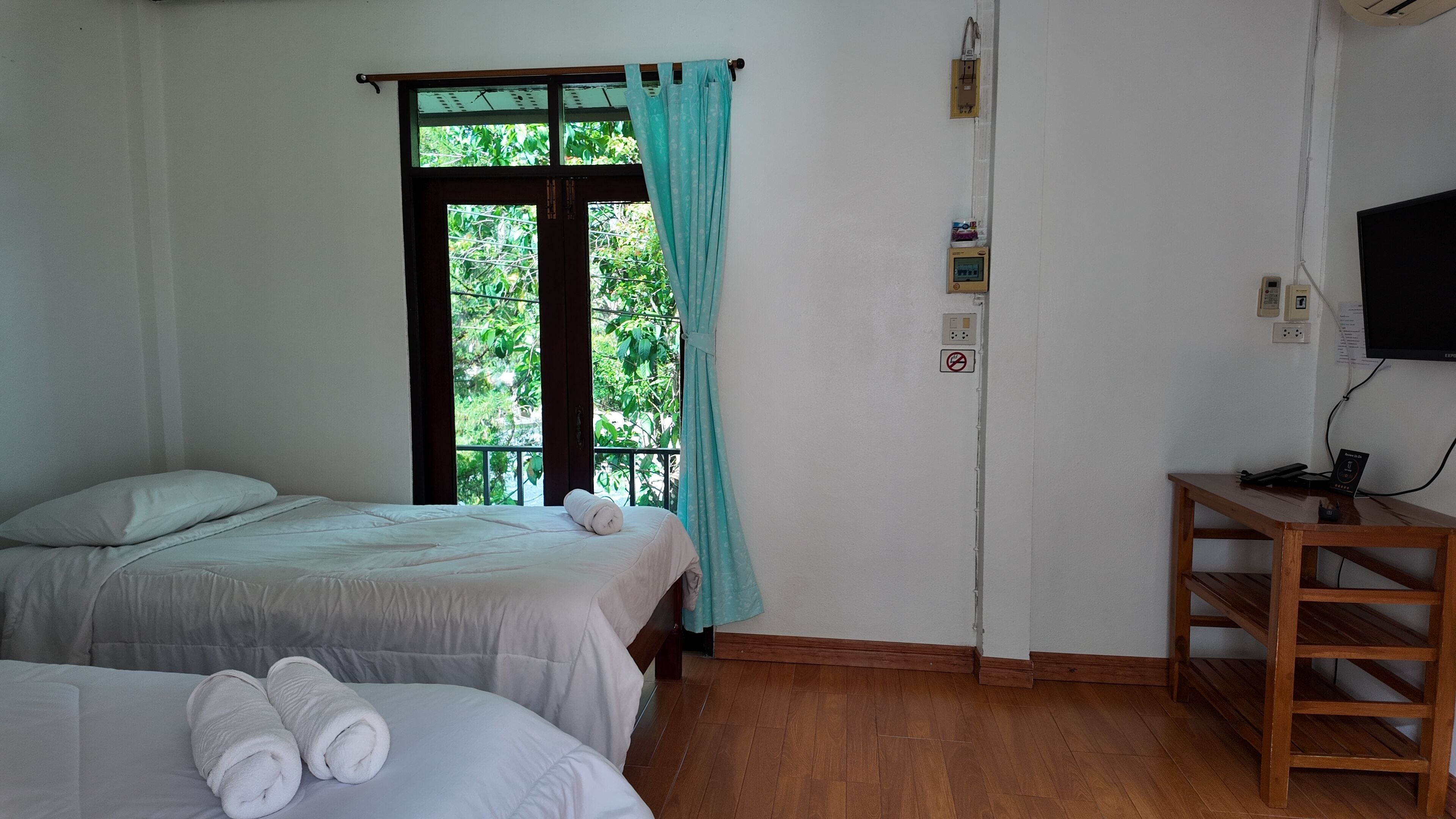 A room with two beds, a wooden floor, a window with curtains, a TV mounted on the wall, and a small table.