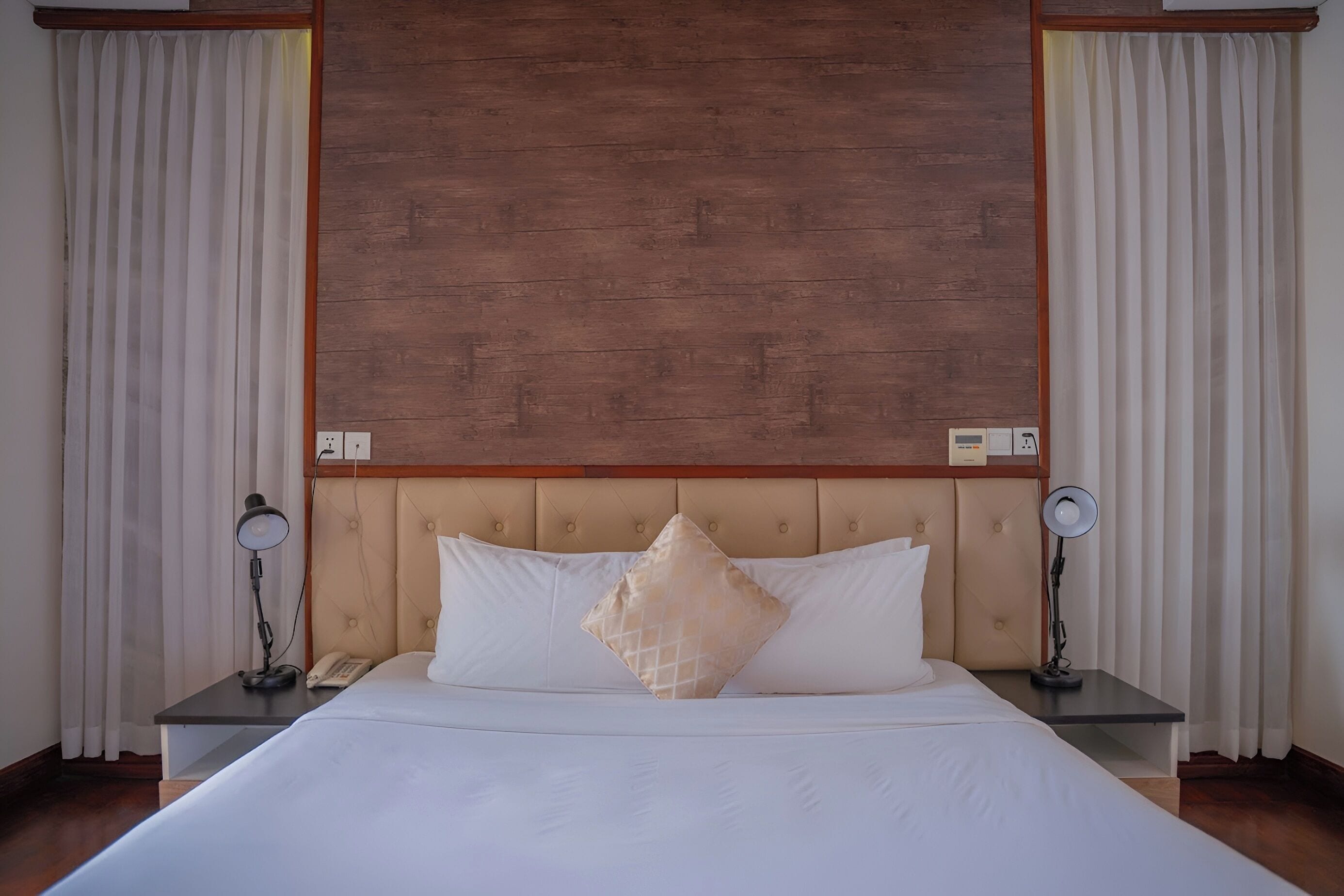 A hotel room with a large bed, two bedside tables with lamps, a wooden headboard, and white curtains.
