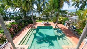 A swimming pool with a view of the ocean, surrounded by palm trees and a patio area with lounge chairs. - Beach House Cairns (Holloways Beach)