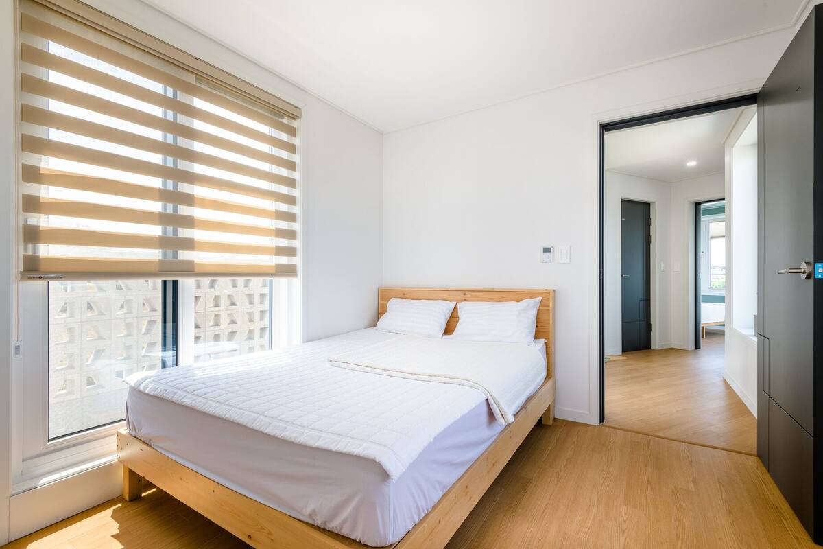 A modern bedroom with a wooden bed, white bedding, and a large window with blinds.
