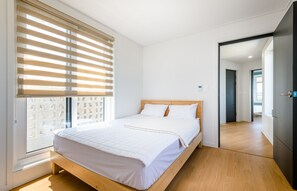 Une chambre moderne avec un lit en bois, une literie blanche et une grande fenêtre équipée de stores.