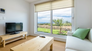 Un salon moderne avec un téléviseur à écran plat posé sur un meuble en bois, une table basse en bois, un canapé blanc agrémenté d’un coussin vert et une grande fenêtre avec des stores donnant sur un paysage.