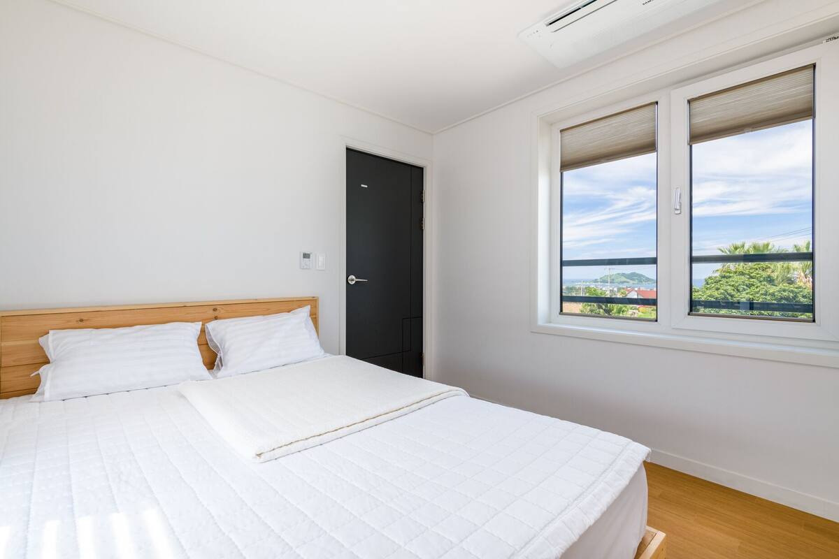 A bedroom with a bed, a black door, and a window with a view of greenery and buildings.