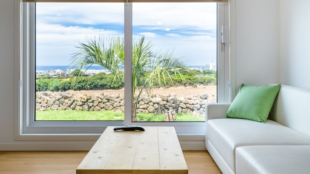 Une pièce avec une grande fenêtre, un canapé blanc orné d’un coussin vert et une table en bois avec une télécommande.