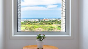 Une fenêtre donnant sur un paysage urbain côtier, une plante en pot posée sur une table en bois et un mur blanc.