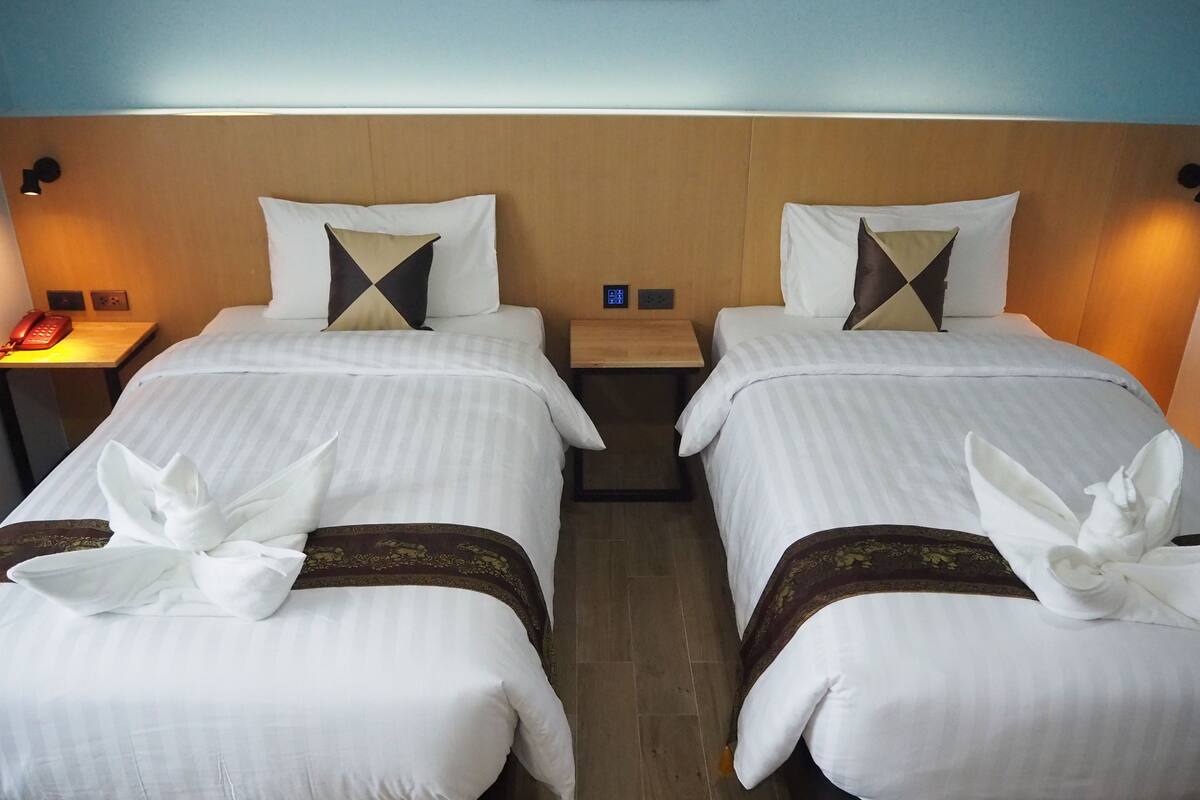 Two neatly made beds with white linens and decorative pillows, a bedside table with a lamp, and a wall-mounted control panel.