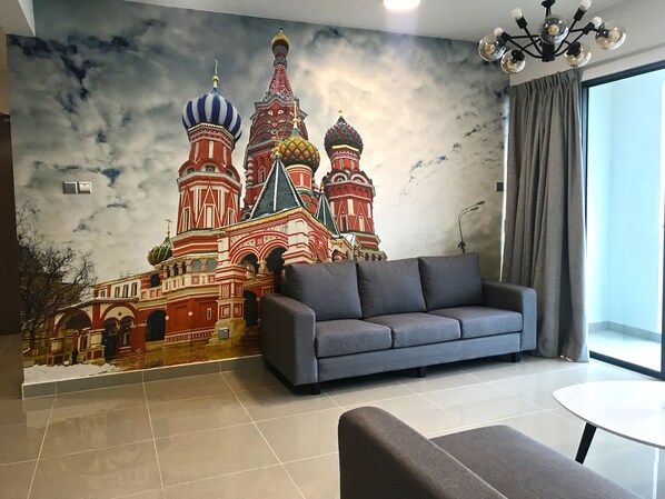 A living room with a large mural of Saint Basil's Cathedral, a grey sofa, a small table, and a view of the outside through a window with curtains.