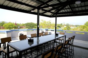 Un área de comedor al aire libre con una mesa larga y sillas, techo cubierto y vistas a zonas verdes y colinas.