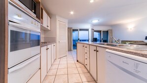 Een moderne keuken met witte apparaten, een inbouwoven en een vaatwasser. Er is een groot raam en een deur die leidt naar een balkon.