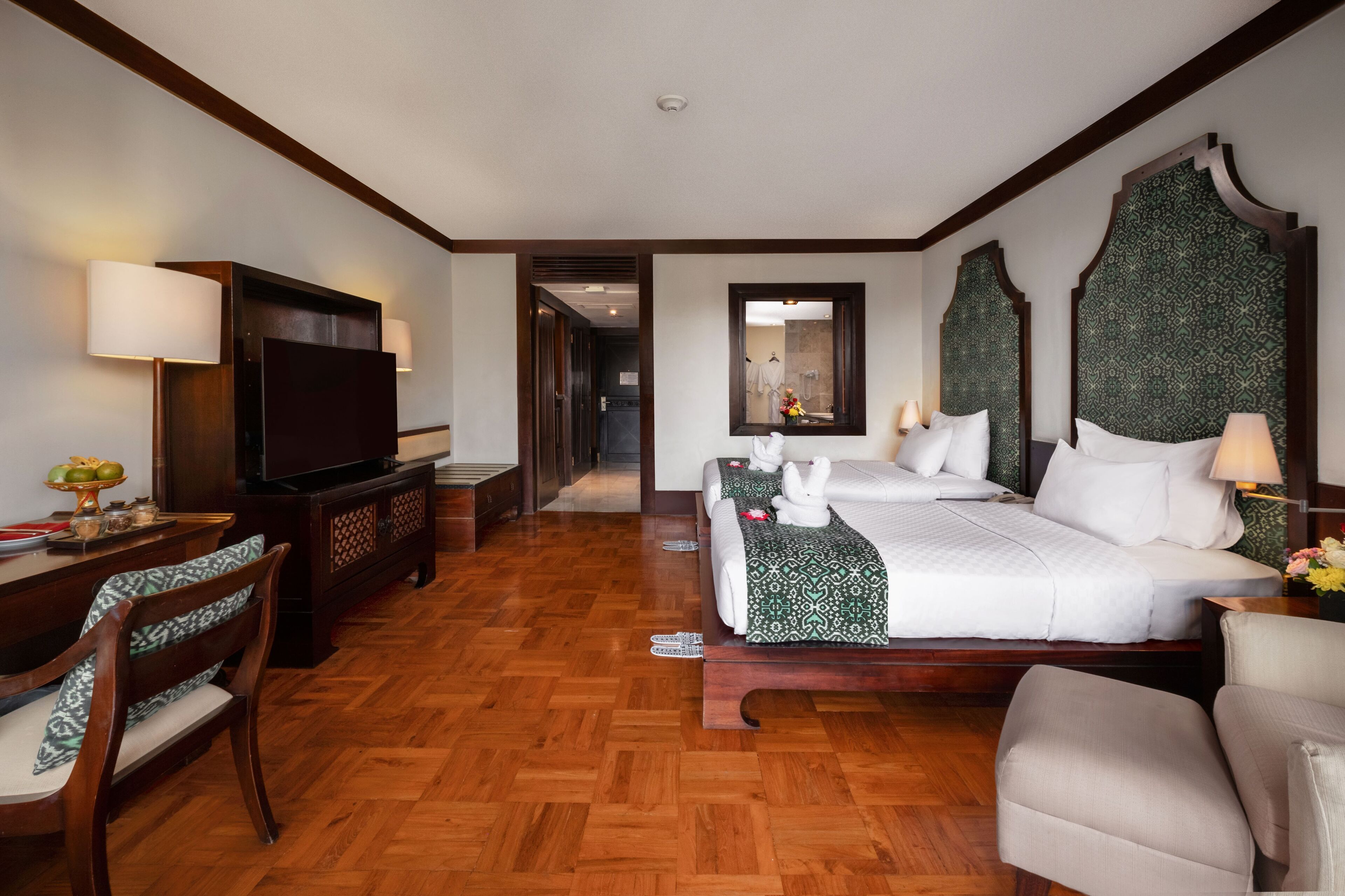 A hotel room with a wooden floor, a large bed with white bedding, a TV, a desk with a lamp, and a chair.