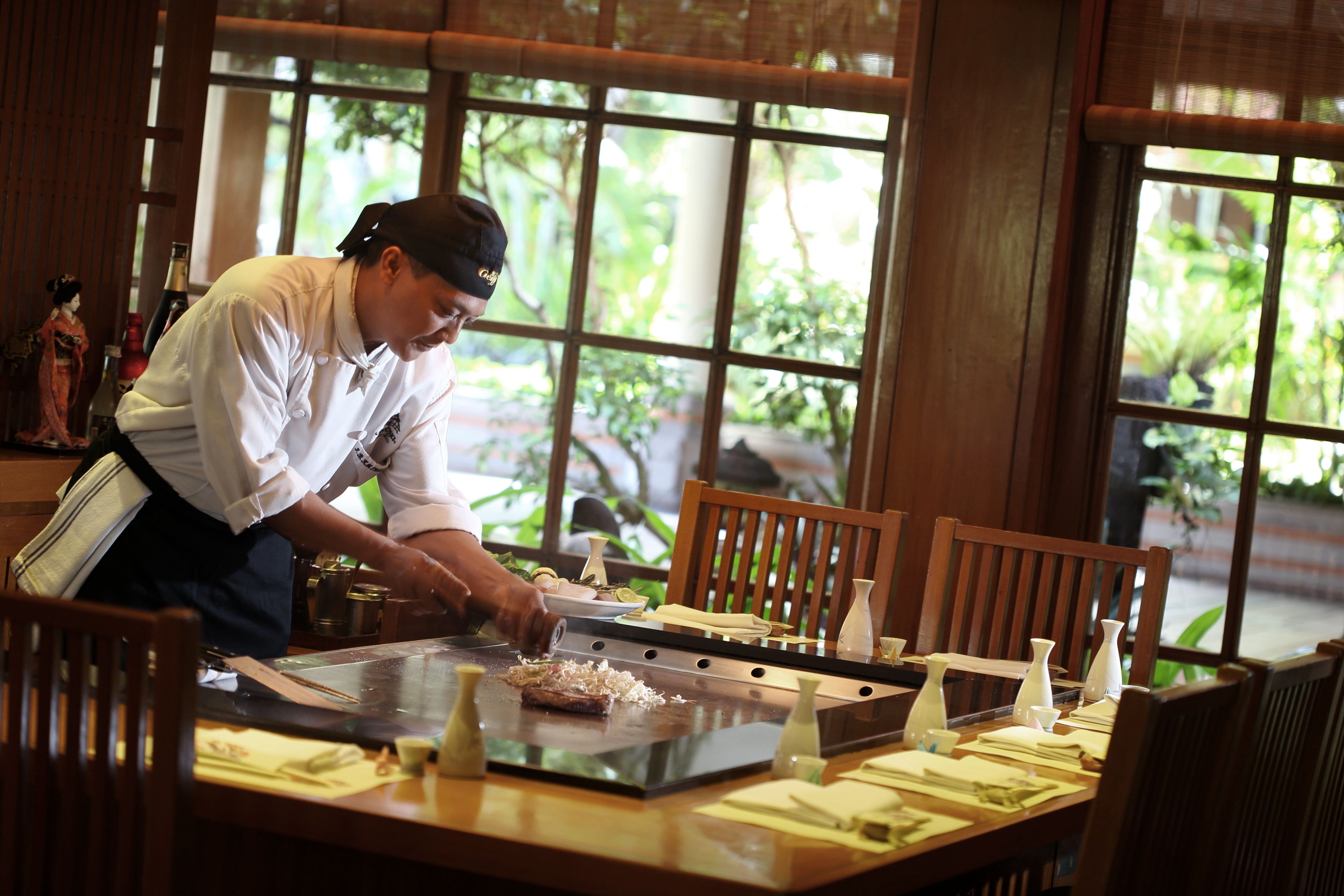A chef preparing food at a wooden table with a view of greenery outside.