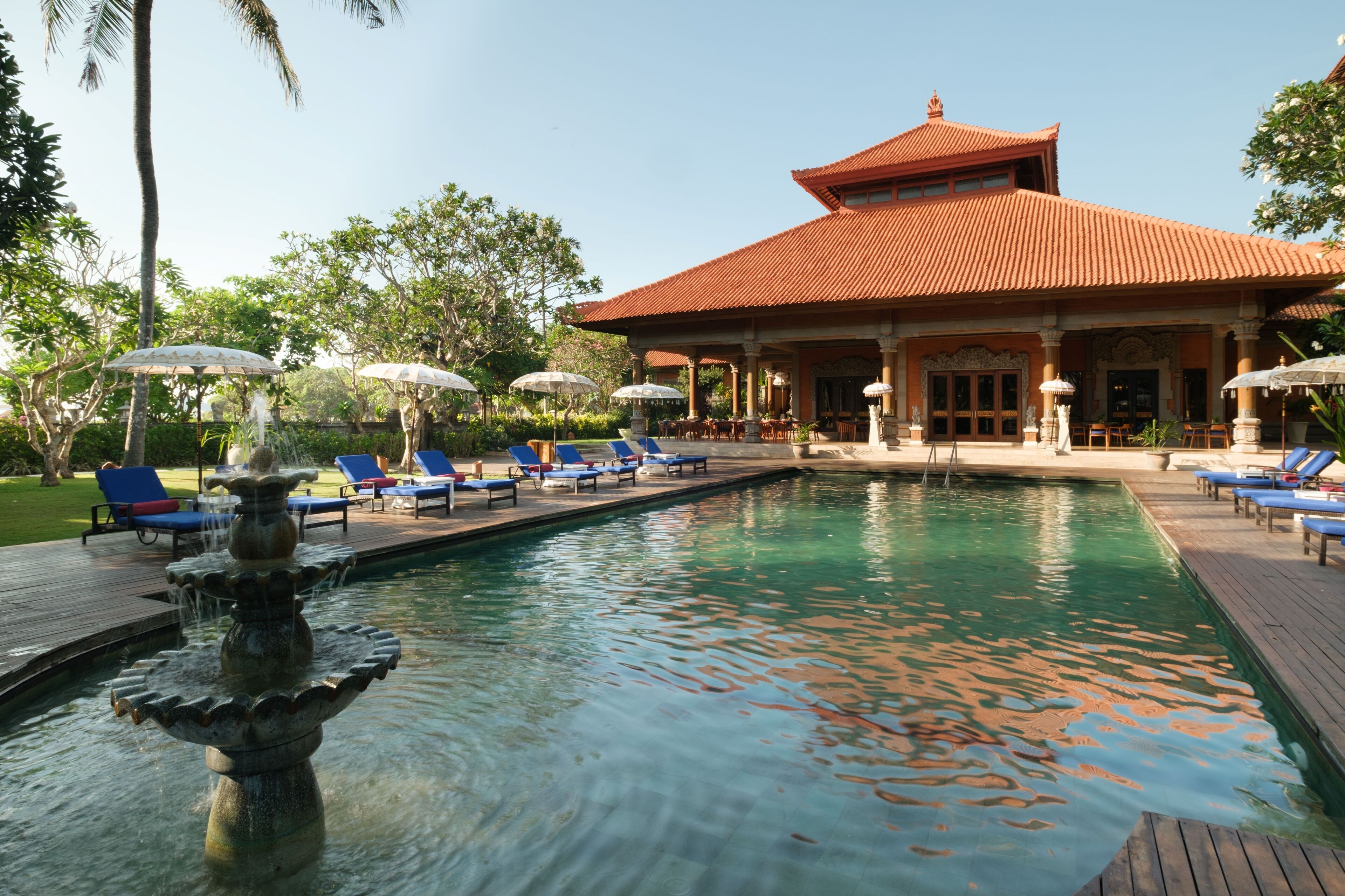 Une piscine avec une fontaine, entourée de chaises longues et de parasols, avec un bâtiment en arrière-plan.
