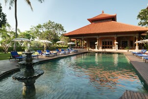 Une piscine avec une fontaine, entourée de chaises longues et de parasols, avec un bâtiment en arrière-plan.