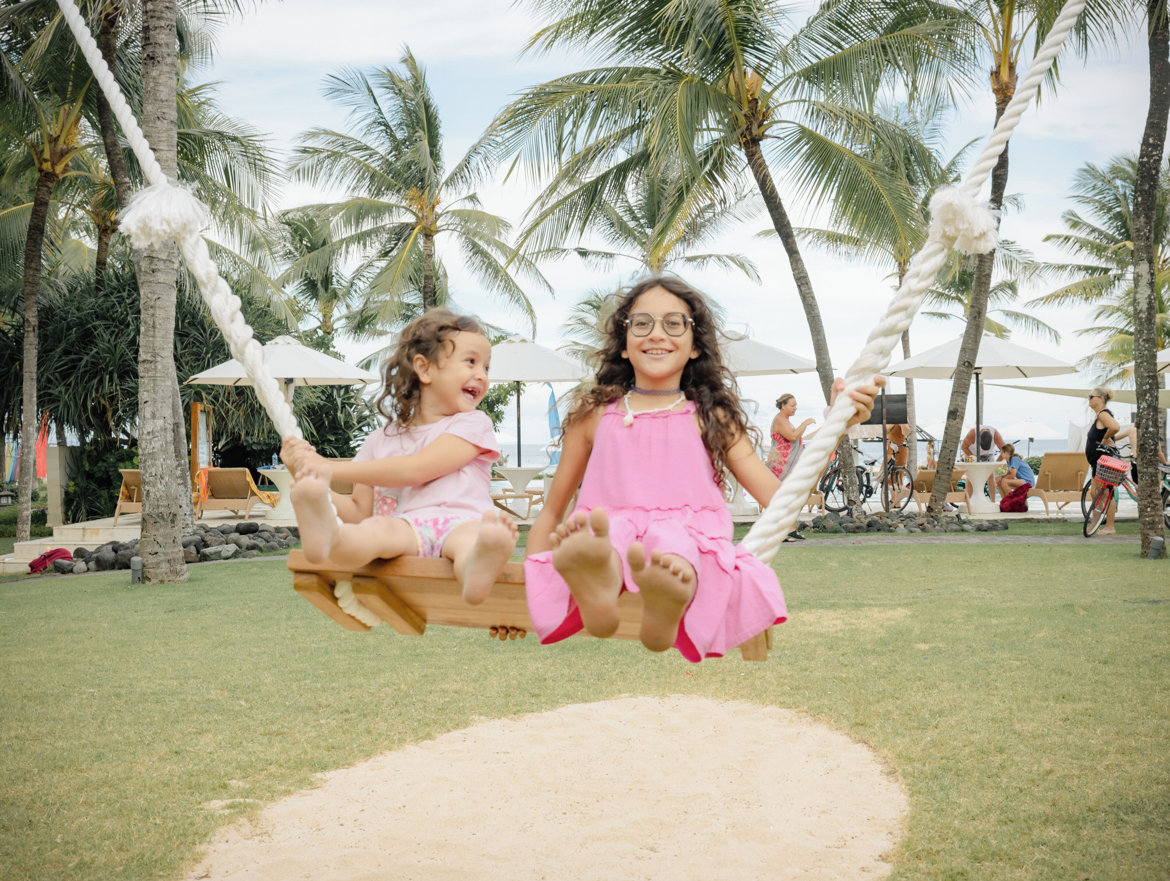 Due bambini su un'altalena in un ambiente tropicale con palme e posti a sedere all'aperto.