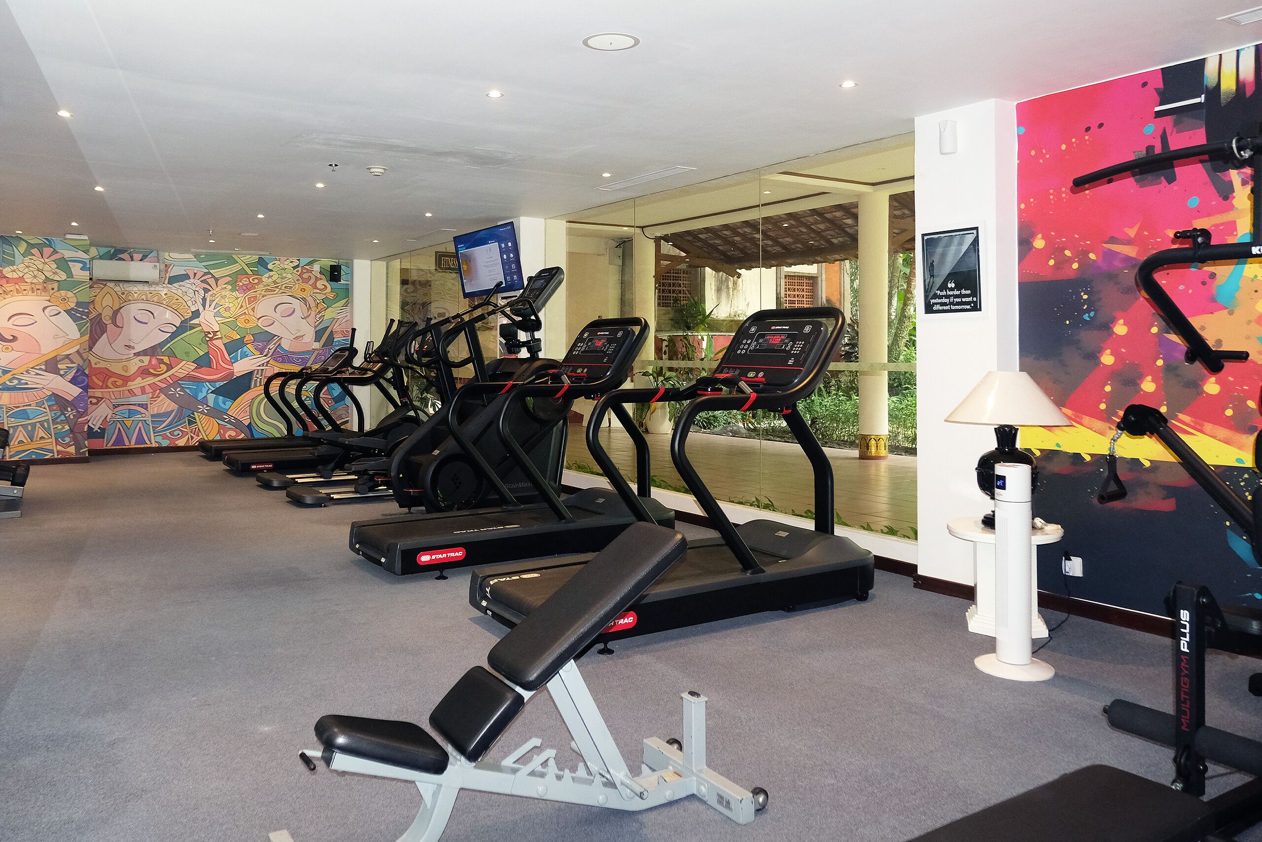 Une salle de sport équipée de tapis de course, d’un banc de développé couché et d’un mur décoré d’œuvres d’art colorées.