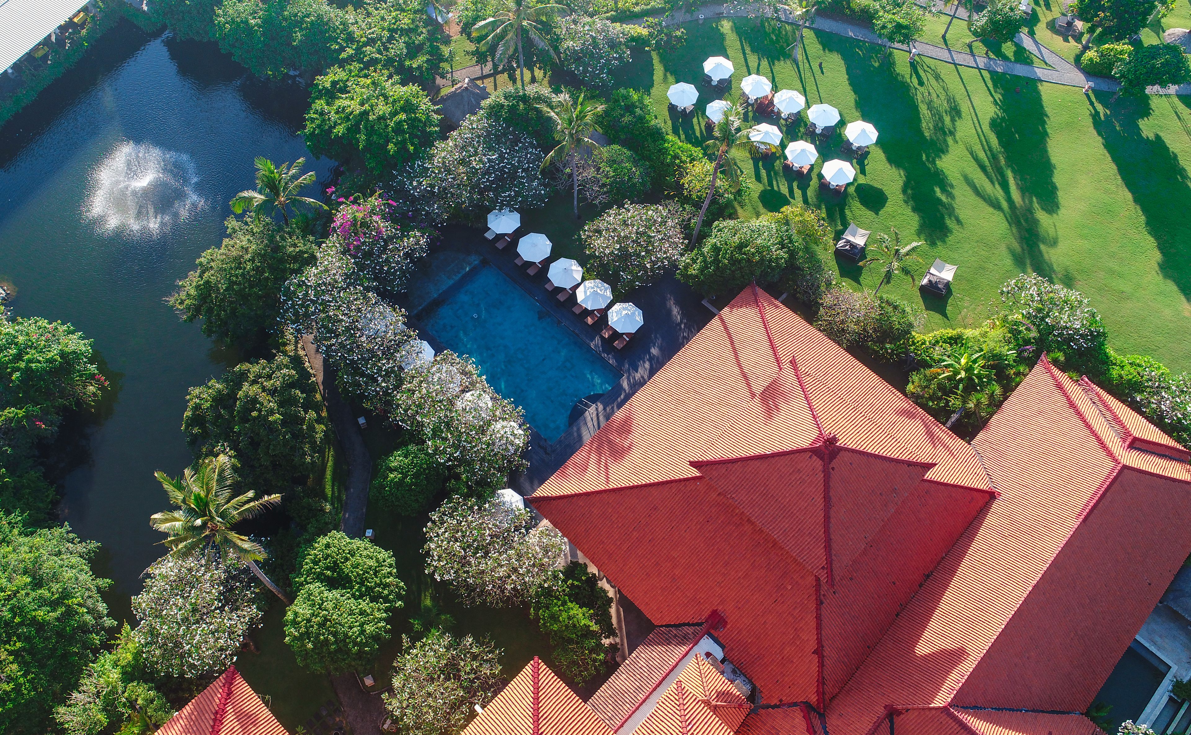 An aerial view of a resort with a swimming pool surrounded by lush greenery, palm trees, and outdoor seating areas with umbrellas.