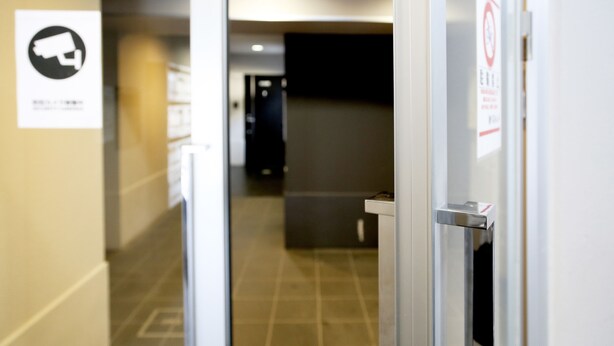 A hallway with a security camera sign, a no-smoking sign, and a door with a handle. - JL Residence Hotel (Osaka)