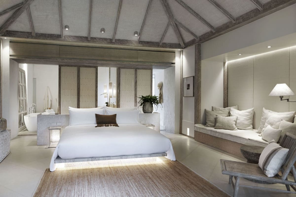 A modern bedroom with a large bed, a seating area with cushions, a side table, and a large mirror.
