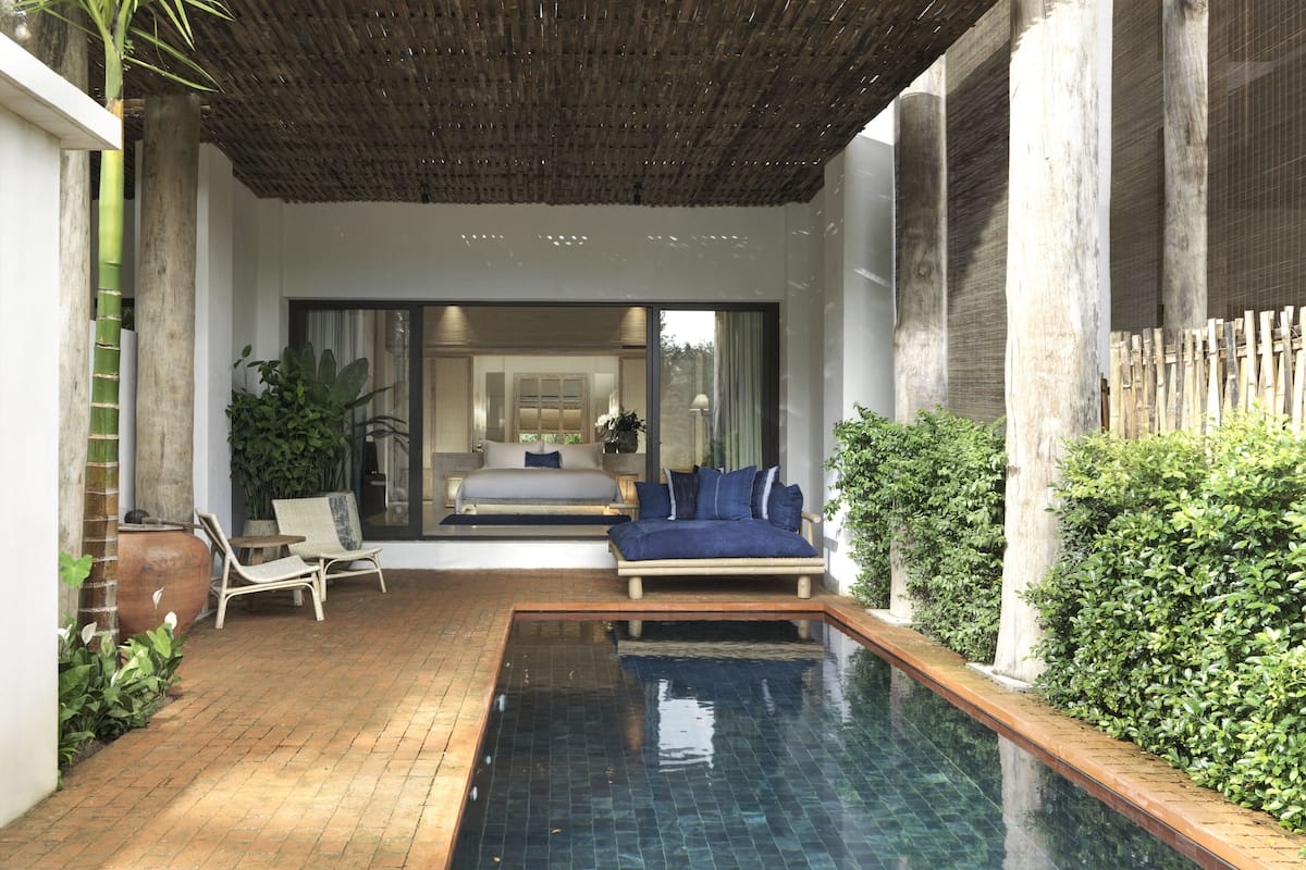 A modern outdoor pool area with a seating area, a view into a bedroom, and a bamboo fence.