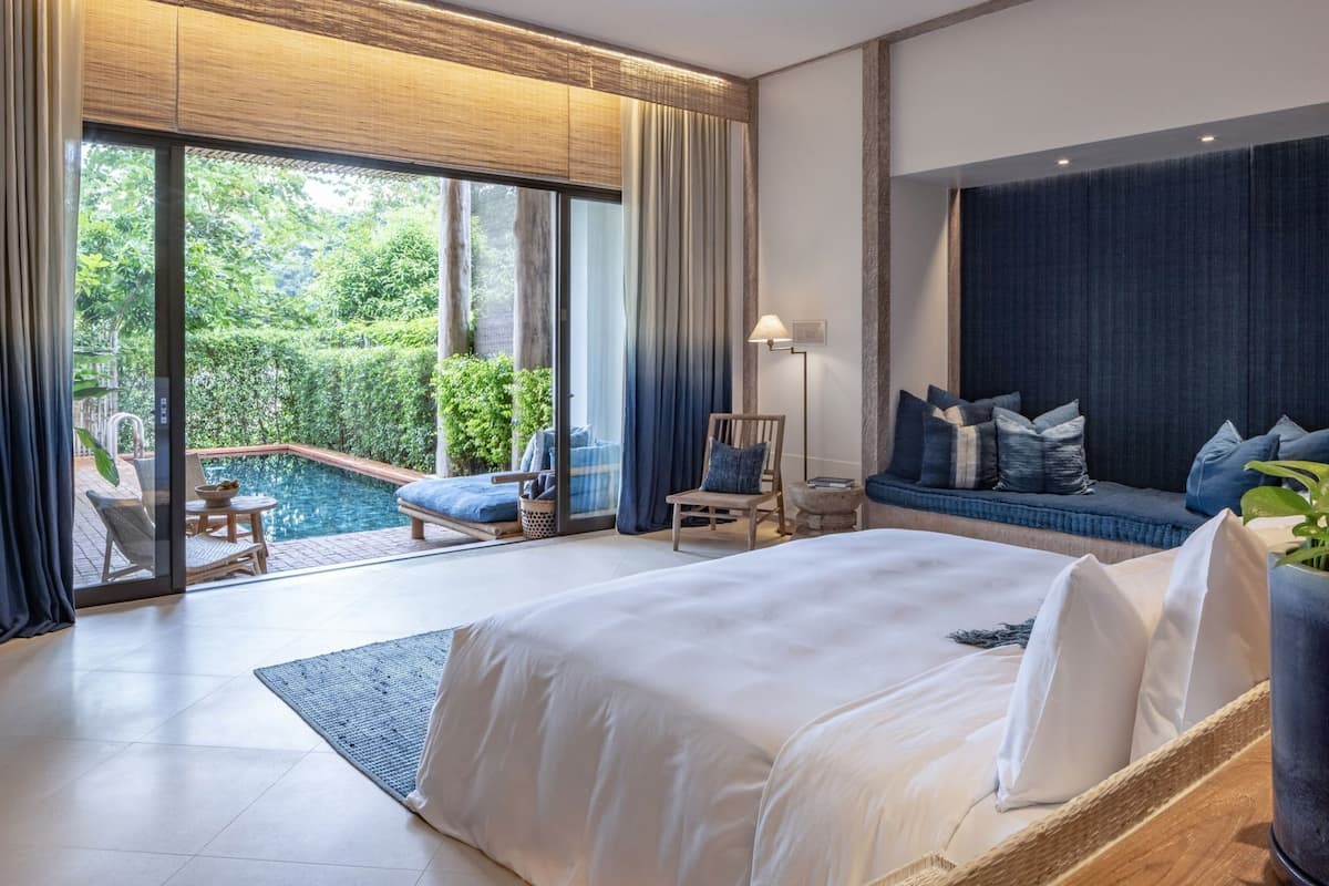 A modern hotel room with a large bed, a seating area with a view of a pool, and a balcony with lush greenery.