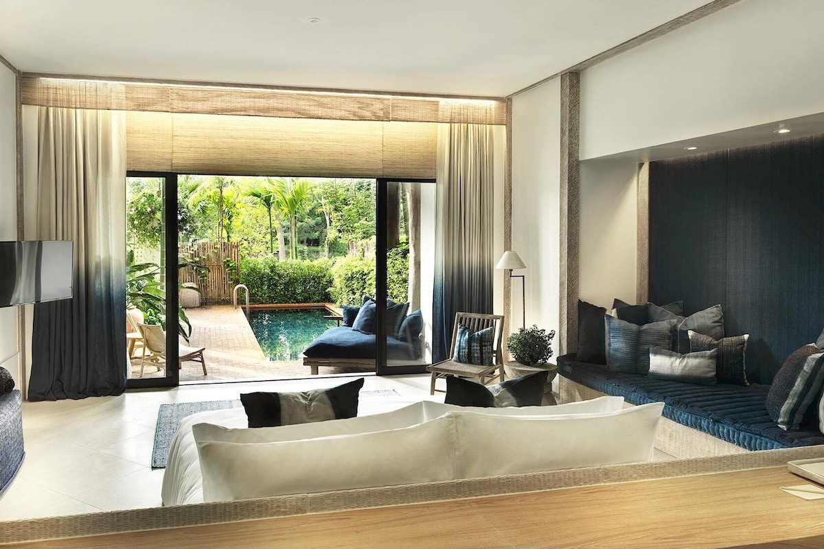 A modern hotel room with a large sliding glass door leading to a pool area, a sofa, a TV, and a bedside table.