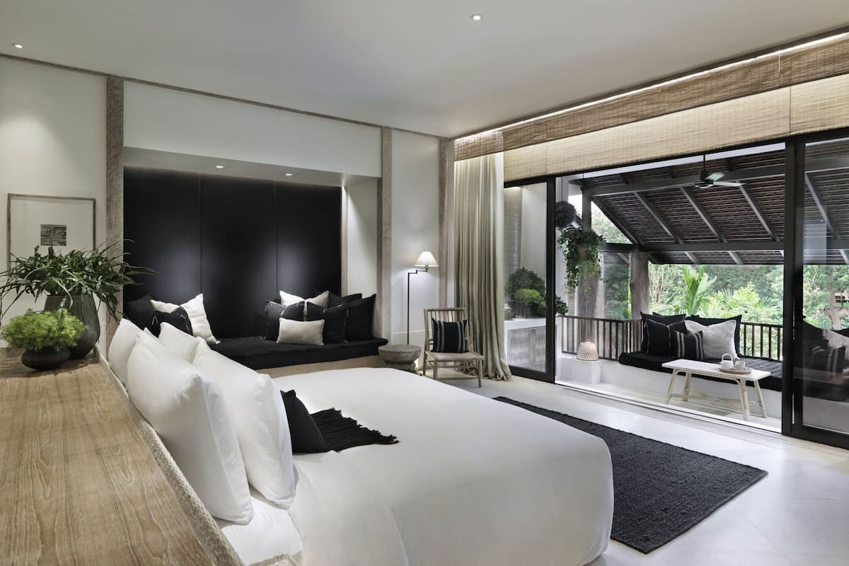 A modern bedroom with a large bed, a seating area with a sofa and chairs, a balcony with plants, and a view of greenery.