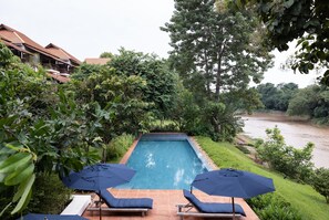 A rectangular pool with two lounge chairs and umbrellas, surrounded by lush greenery and overlooking a river.