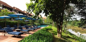 A poolside area with lounge chairs and umbrellas, a river, and a building in the background.
