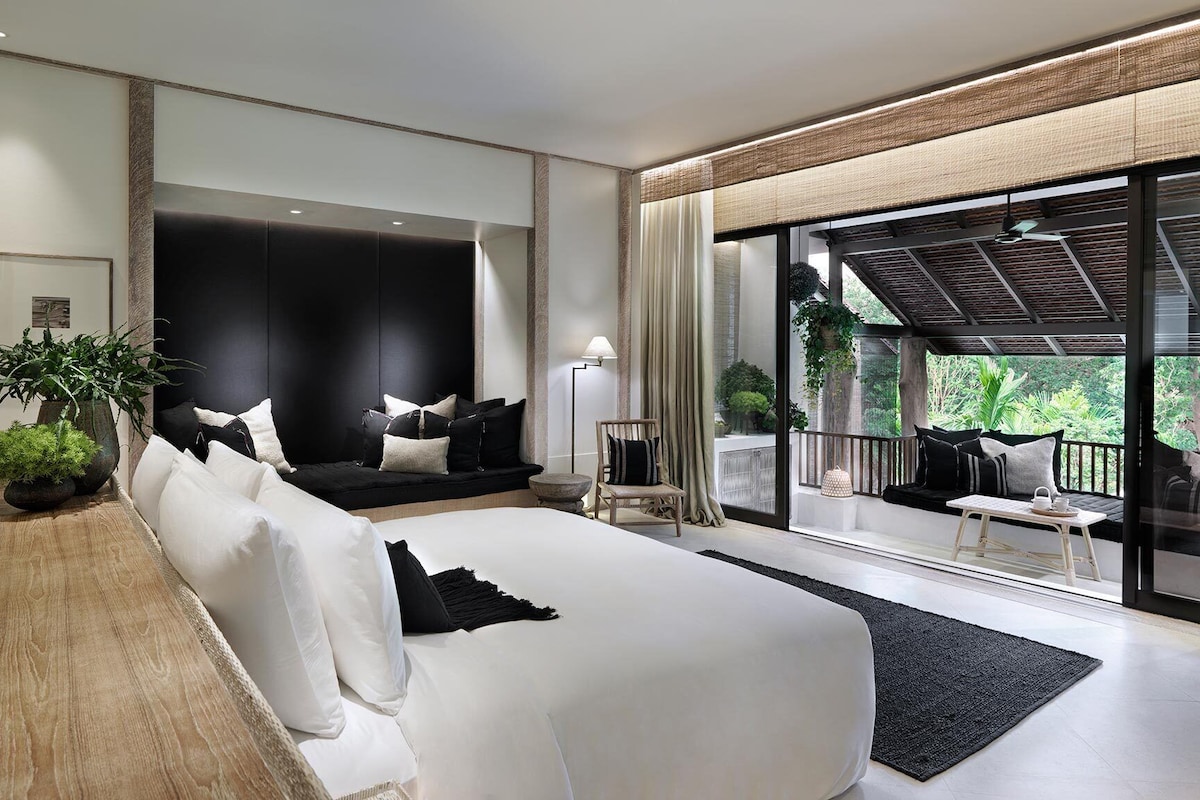 A modern bedroom with a large bed, a seating area with a sofa and chairs, and a balcony with plants.