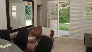A room with a brown leather sofa, a dining table with brochures, a large mirror, and a sliding glass door leading to a garden. - Umhlanga Lodge -No children (Umhlanga)