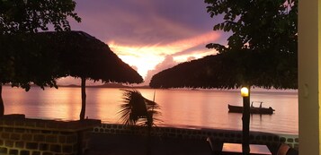 Vista al atardecer desde una terraza con paredes de piedra, palmeras y un barco en el agua.