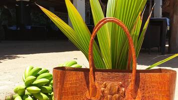 A large, textured brown bag with a handle, placed on sandy ground next to a bunch of green bananas and a plant with long green leaves.