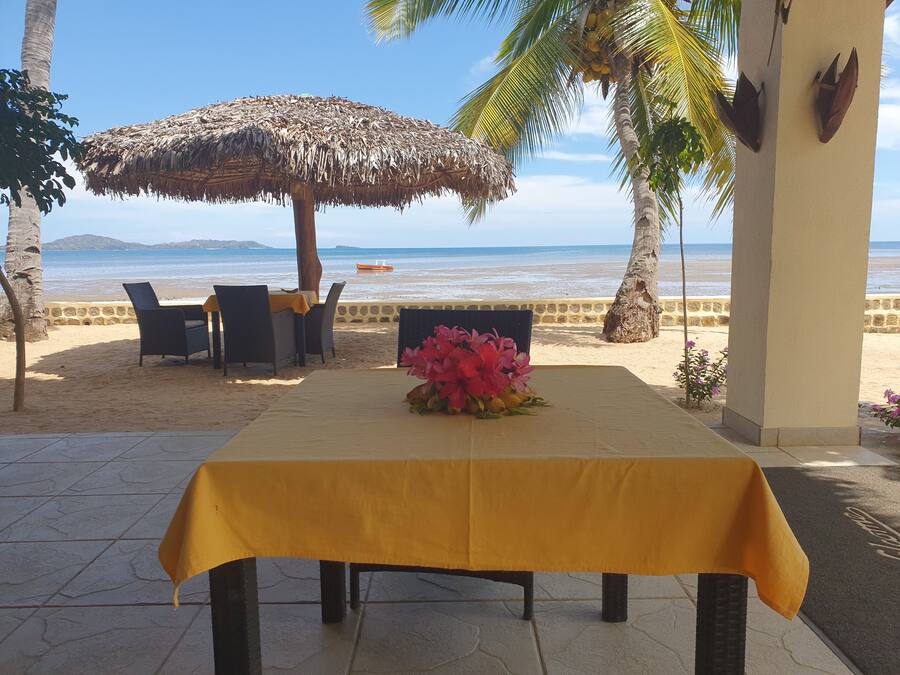 A beachside table with a yellow tablecloth, surrounded by wicker chairs, a thatched umbrella, and a flower centerpiece.