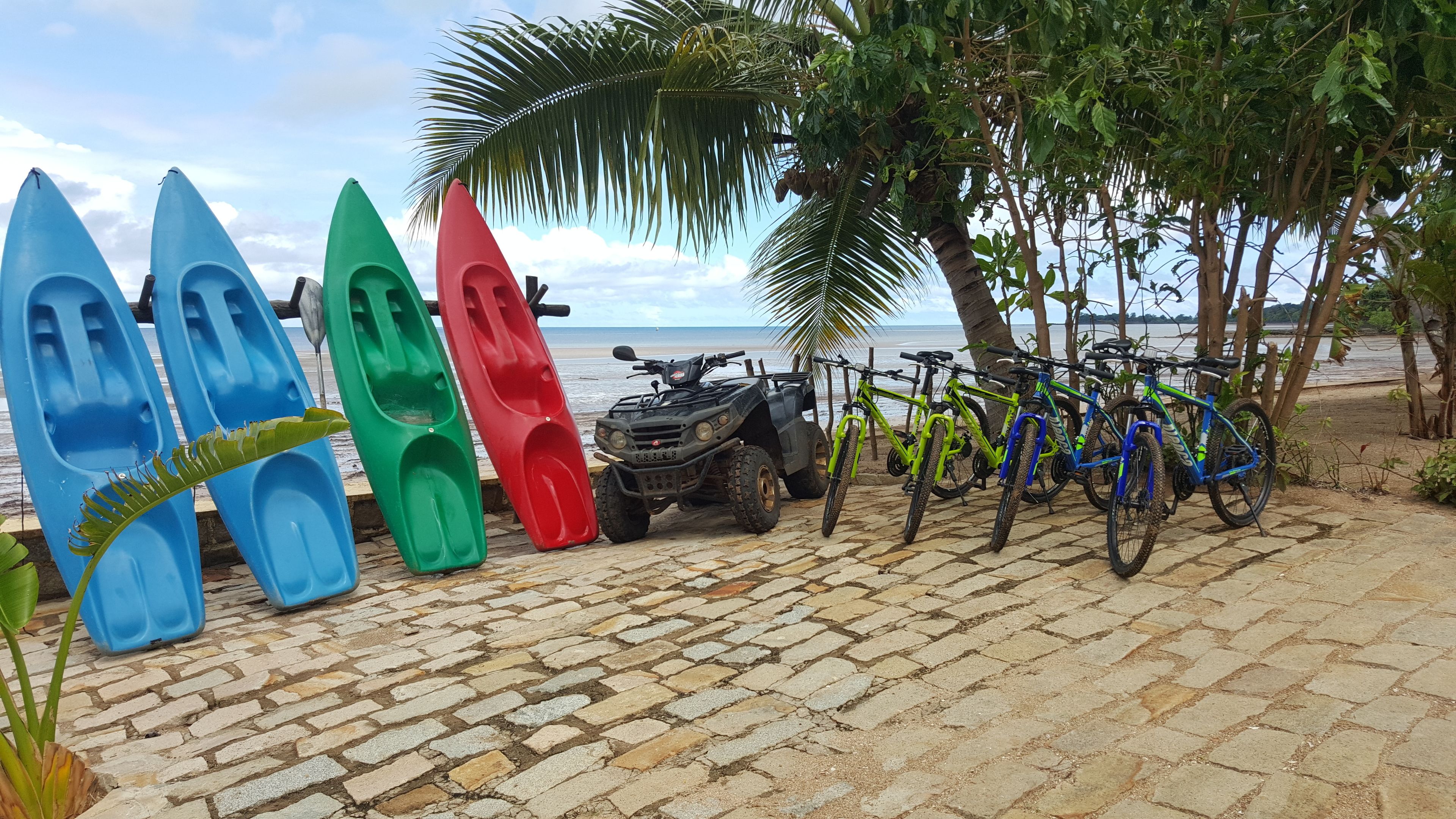 Kayaks, an ATV, and bicycles parked on a cobblestone path by the beach.